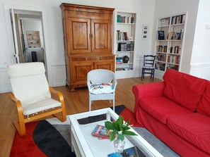 A living room with a white armchair, a red sofa, a coffee table with magazines and a vase, a wooden cabinet, and a bookshelf. - Apartment Quimper hyper-centre, 3 bedrooms (Quimper)