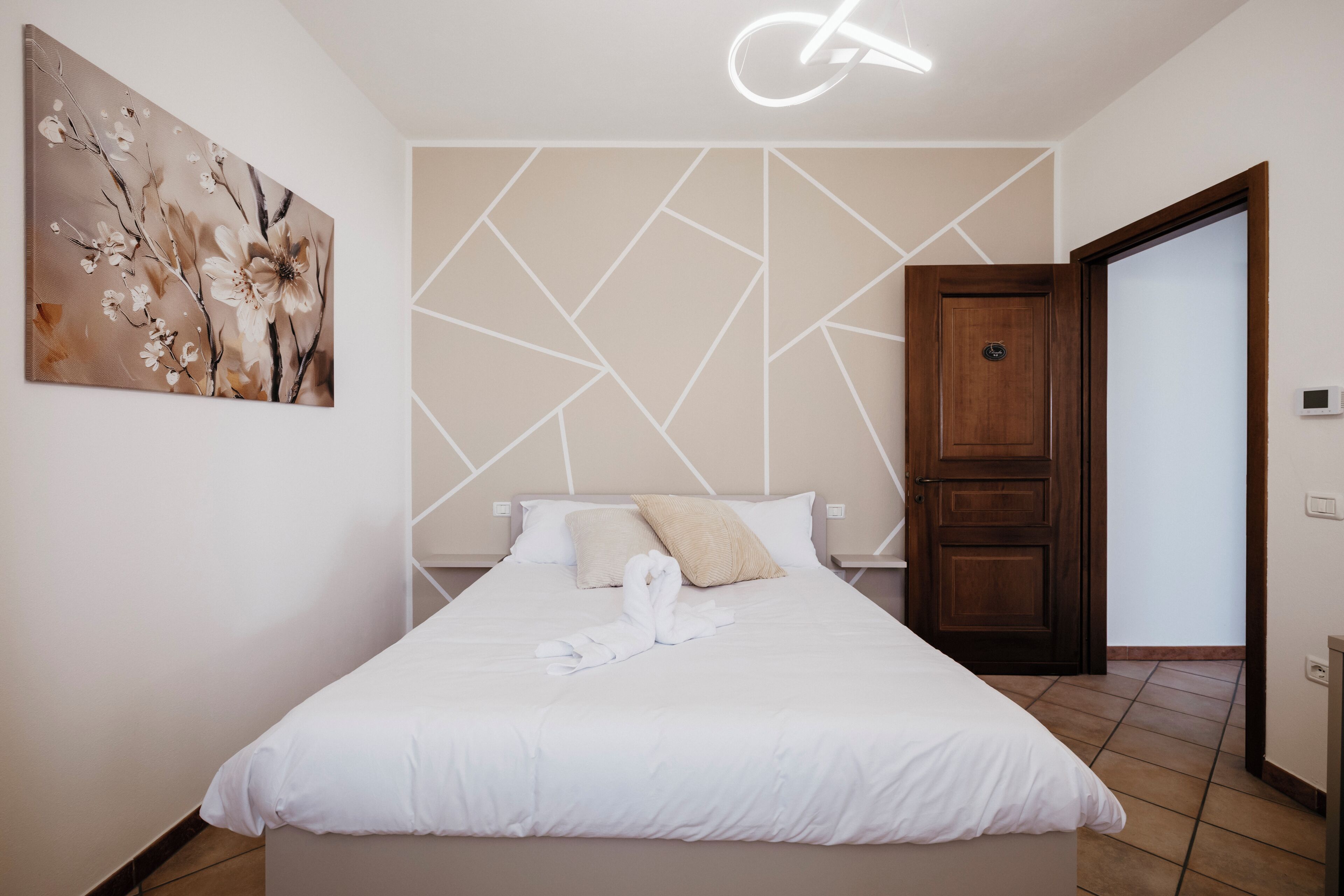 Un lit bien fait, avec du linge de lit blanc et une serviette, un mur orné d’un motif géométrique et un tableau représentant des fleurs.