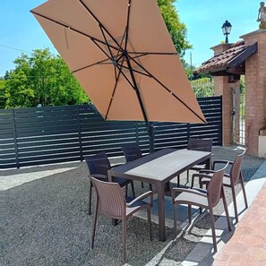 An outdoor patio with a table and chairs, a large umbrella, and a brick building in the background. - Private Relax Apartment: Sauna and Mini Pool (Piemonte)