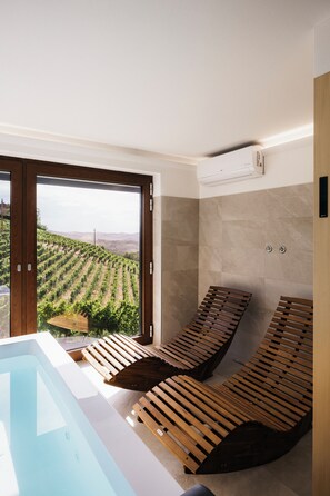 A spa area with a hot tub, wooden loungers, and a view of a vineyard through a large window. - Private Relax Apartment: Sauna and Mini Pool (Piemonte)