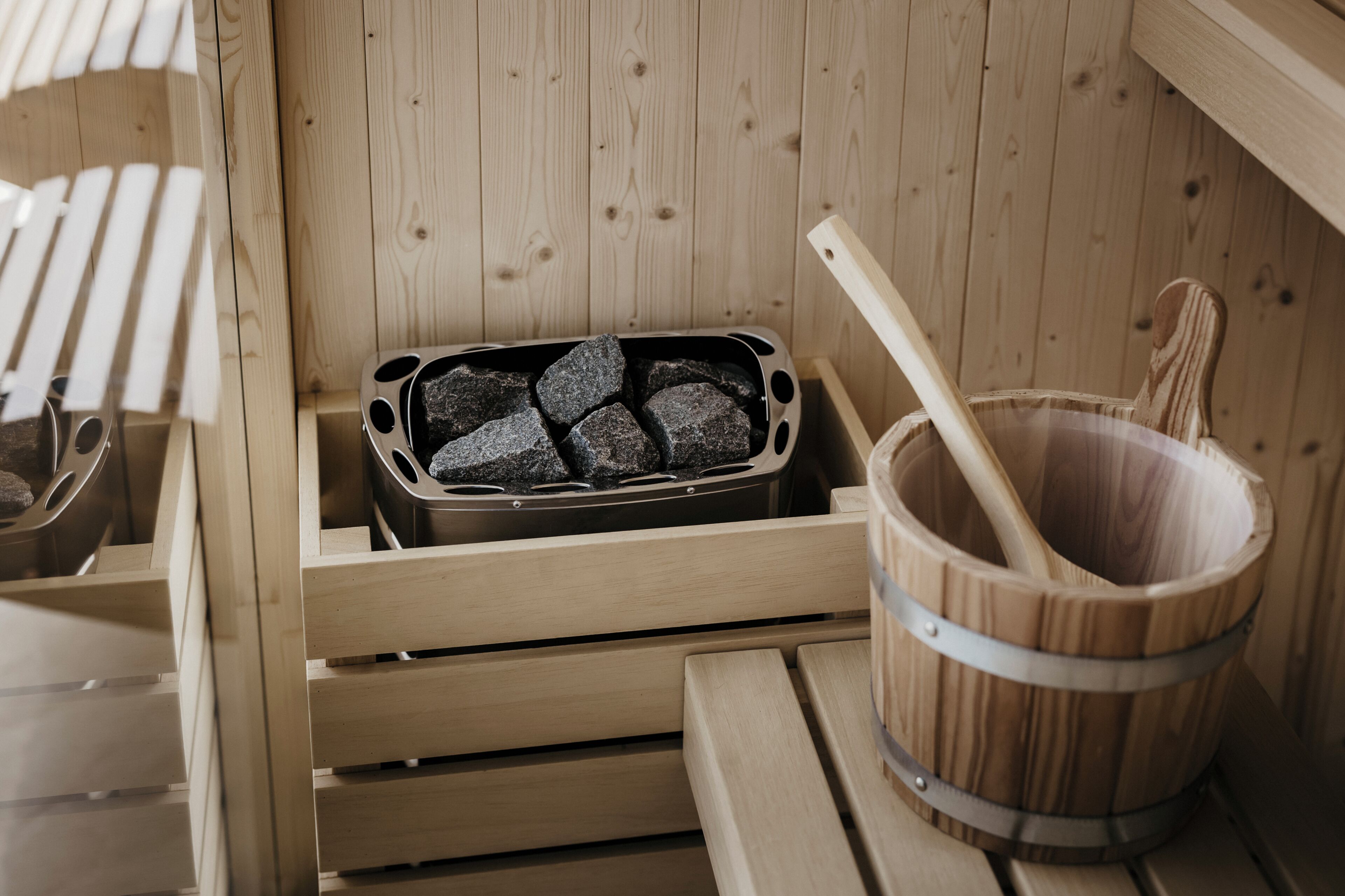 A sauna with a wooden bench, a metal container with stones, and a wooden bucket with a handle.