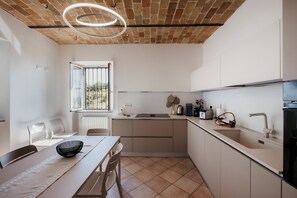 A modern kitchen with a brick ceiling, white cabinetry, a dining table with chairs, and a window with a view of greenery. - Private Relax Apartment: Sauna and Mini Pool (Piemonte)