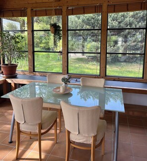 Uma área de refeições com mesa de vidro, quatro cadeiras, uma planta em vaso e um vaso com flores.