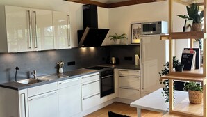 A modern kitchen with white cabinets, a black stove, and a wooden dining table with chairs. - Fantastic vacation home close to nature with sauna (Amelinghausen)