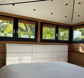 Quarto com cama grande, cabeceira de madeira e três janelas com persianas.