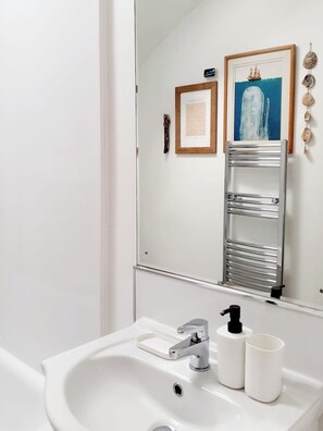 Un baño con un lavabo blanco, grifo cromado y un espejo que refleja un cuadro enmarco y un perchero.
