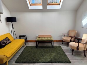 Una habitación con sofá amarillo, un otomán verde, dos sillones, una lámpara de pie y un tragaluz.