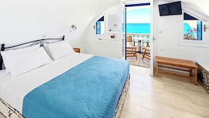 Um quarto com cama, um banco de madeira, uma varanda com vista para o mar e uma janela com vista para um prédio.