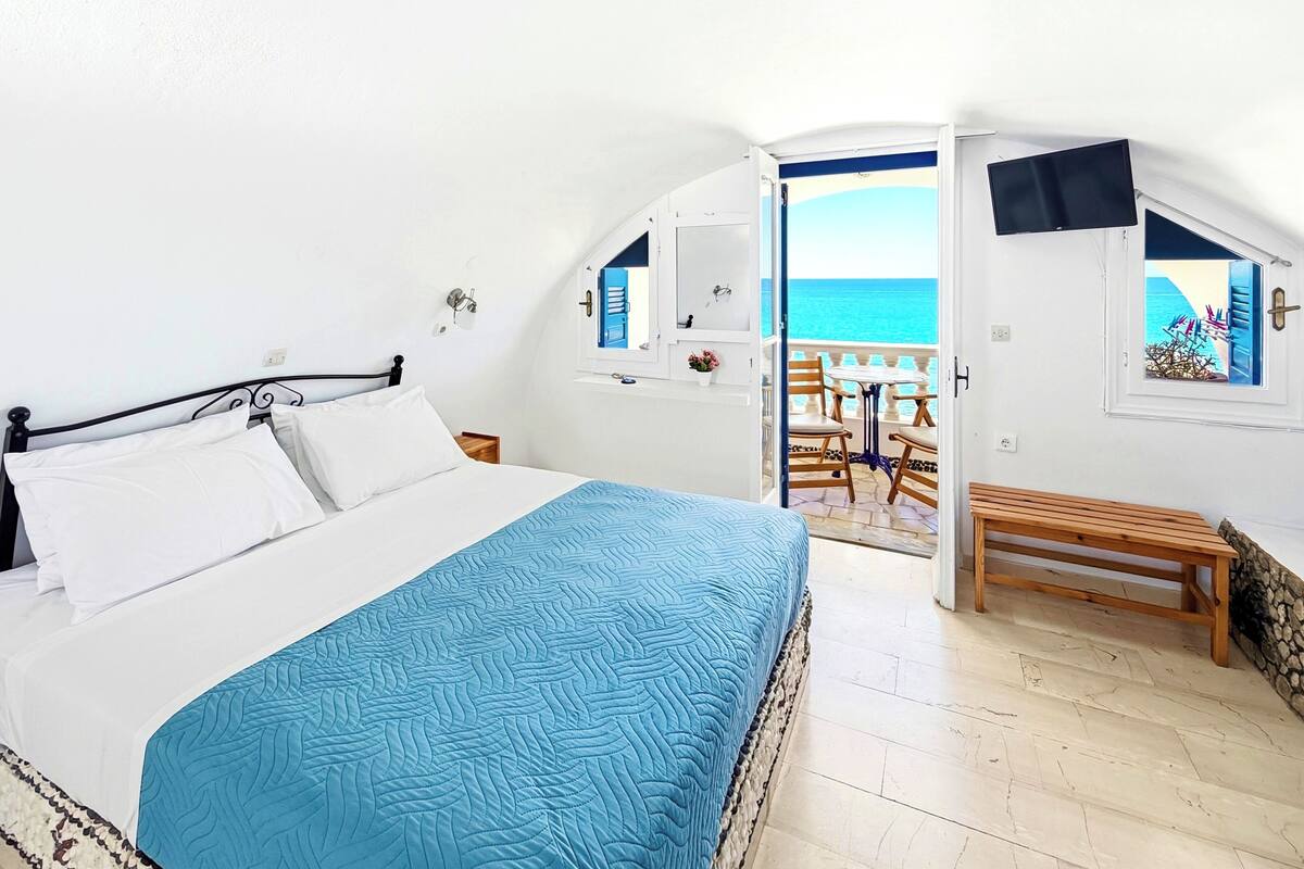 Un dormitorio con cama, un banco de madera, un balcón con vista al mar y una ventana con vista a un edificio.