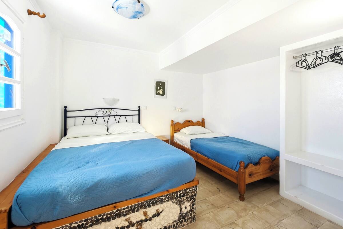 Una habitación con dos camas, un ventanal pequeño, un estante en la pared y una puerta con perchero.