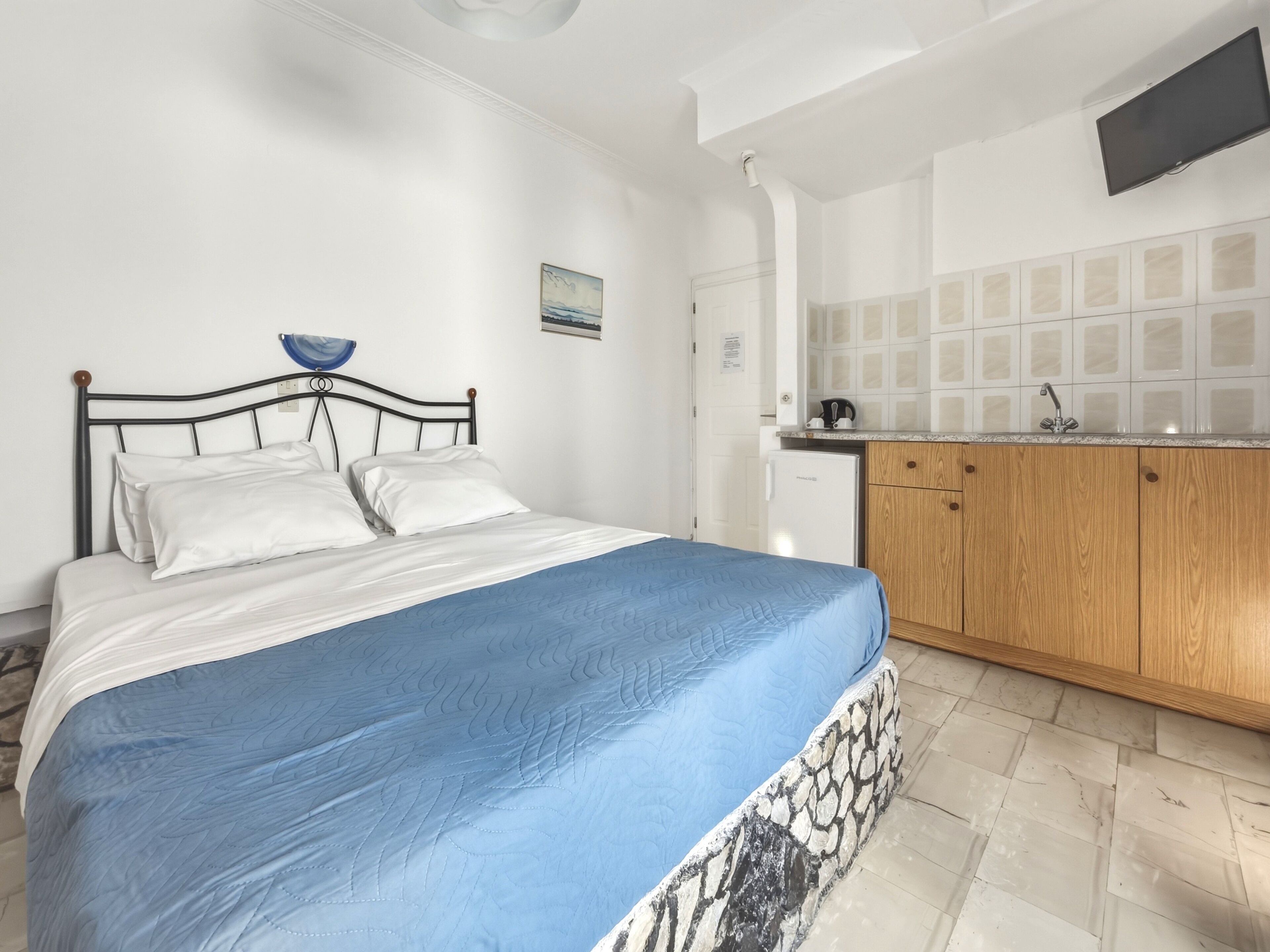 Ein Schlafzimmer mit einem Bett, einer blauen Tagesdecke, einer weißen Wand, einer kleinen Küchenzeile inklusive Kühlschrank, Holzschränken und einer gefliesten Rückwand.