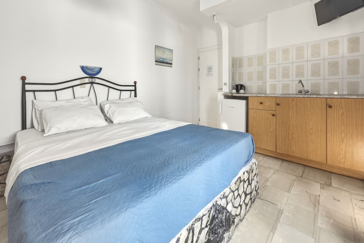 A bedroom with a bed, a blue bedspread, a white wall, a small kitchen area with a refrigerator, wooden cabinets, and a tiled backsplash.