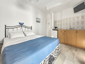 Ein Schlafzimmer mit einem Bett, einer blauen Tagesdecke, einer weißen Wand, einer kleinen Küchenzeile inklusive Kühlschrank, Holzschränken und einer gefliesten Rückwand.