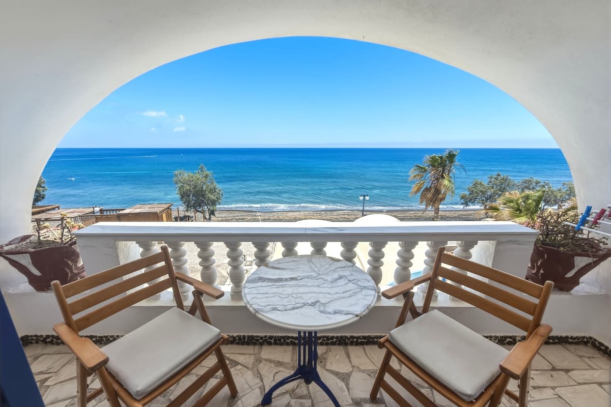 A balcony with two wooden chairs and a marble table, overlooking a beach with the sea and palm trees.