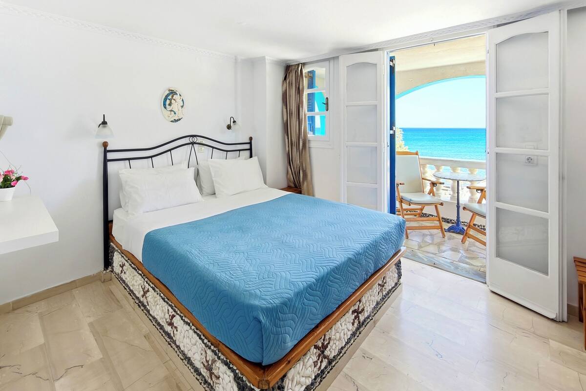 Un dormitorio con cama, mesita de noche, planta en maceta, vista al mar y balcón con mobiliario exterior.