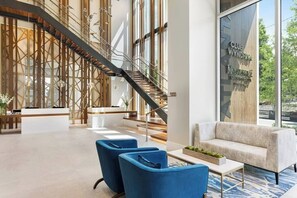 Eine moderne Hotellobby mit einer Rezeption, blauen Sesseln, einer Treppe und einem großen Fenster mit Blick ins Freie.