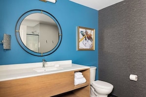 Ein Badezimmer mit einem runden Spiegel, einem weißen Waschbecken, einer Holz-Waschkommode, einer Toilette und einem gerahmten Kunstwerk an der Wand.