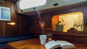 Een houten bank met een boek en een kopje, een spiegel die een plant en een raam reflecteert, en een kleine kaars op de bank.