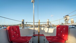 Een dek van een boot met twee rode, beklede zitplaatsen, een stuurwiel en uitzicht op een jachthaven met andere boten.