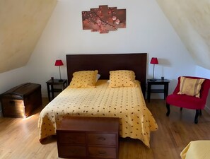 Une chambre Ă coucher comprenant un lit, deux tables de chevet, un fauteuil rouge, une commode en bois et une Ćuvre dâart accrochĂ©e au mur.