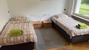 A bedroom with two beds, a gray rug, bedside tables, a lamp, and a window with a view of greenery.