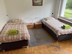 A bedroom with two beds, a gray rug, bedside tables, a lamp, and a window with a view of greenery.