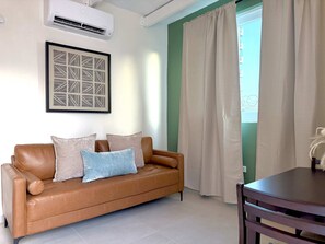 A brown leather sofa with cushions, a framed abstract artwork, an air conditioning unit, and a window with curtains. - Beachside 1BR Apt - Balcony & Pool (San Juan)