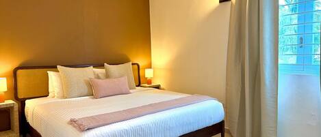Una cama bien hecha con almohadas, una lámpara de noche y una ventana con cortinas.