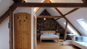 Een slaapkamer met een schuine balkenplafond, houten liggers, een stenen muur, een bed, nachtkastjes en een bank.