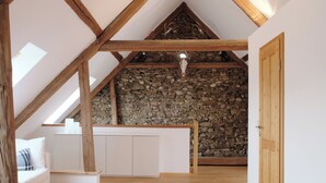 Een loft-achtige kamer met een schuine kap, zichtbare houten balken, een stenen muur en een witte bank.