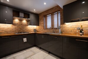 Cozinha moderna com armários escuros, parede de tijolos, fogão a gás e uma garrafa de vinho sobre o balcão.