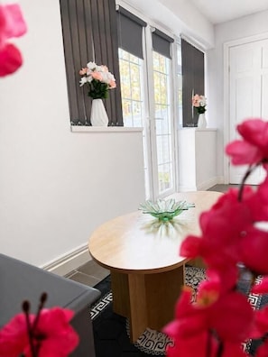 A dining area with a round table, a vase with flowers, and a window with blinds. - RS Prime Apartment for Family Couple  (Solihull)