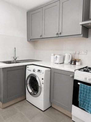 A compact kitchen with grey cabinets, a washing machine, a stove, and a sink. - RS Prime Apartment for Family Couple  (Solihull)