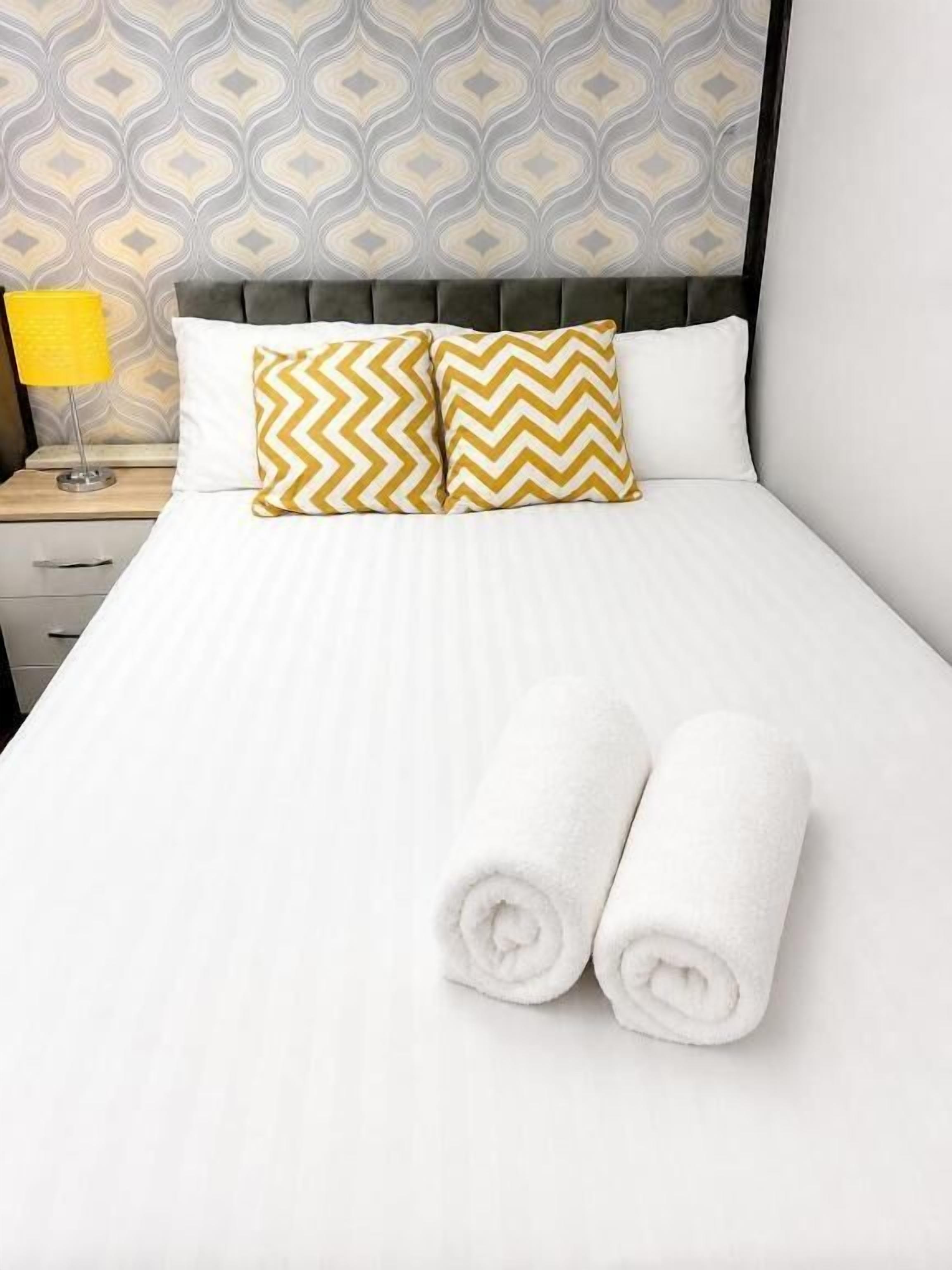 Una cama bien tendida con sábanas blancas, dos almohadas estampadas y toallas enrolladas.