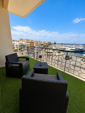 A balcony with wicker furniture, a small table, and a view of a marina with boats and buildings in the background. - HARBOR ELEGANCE ESTATE (Milazzo)