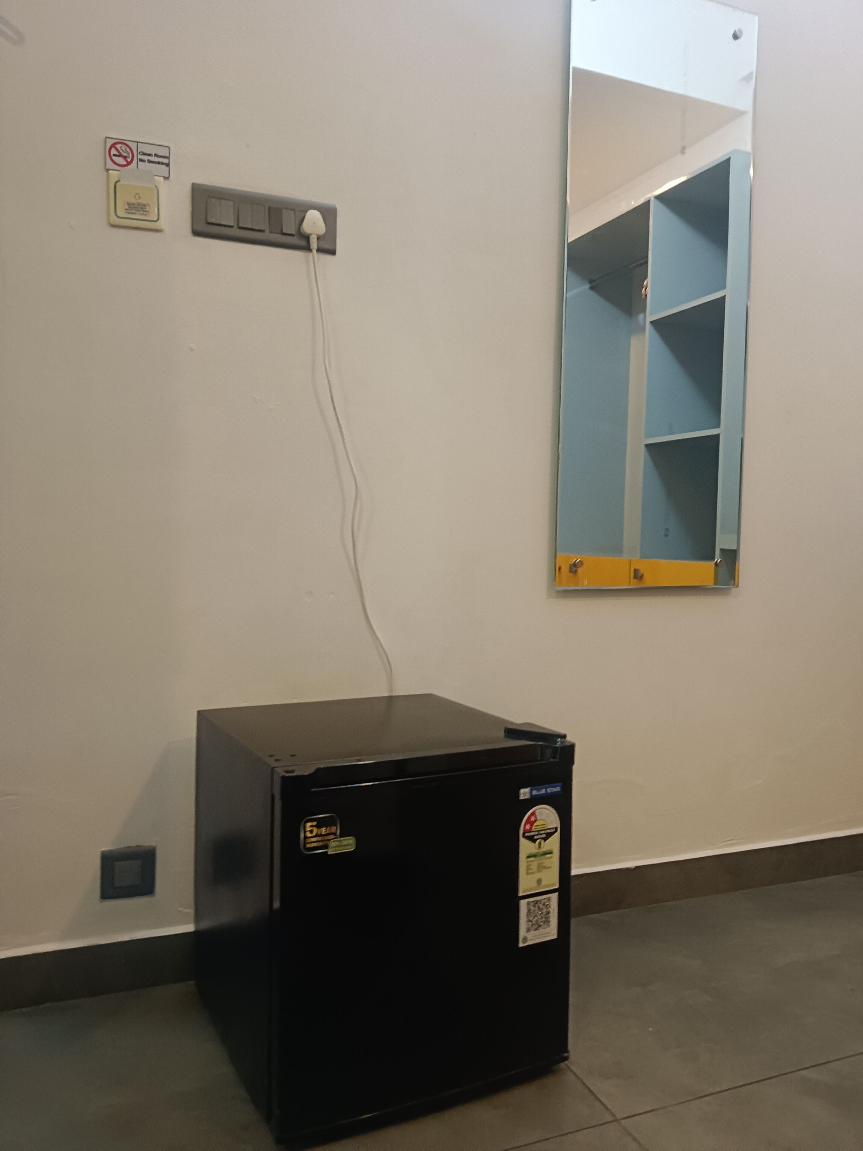 A black refrigerator with a sticker, a power outlet, a no-smoking sign, and a blue shelving unit on a wall.