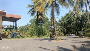 Un espacio al aire libre con pavimento, palmeras, un parque infantil y un edificio con una estructura cubierta.