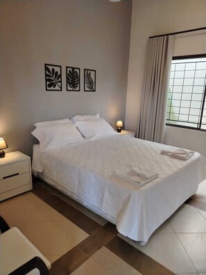 Quarto com cama grande, duas mesas de cabeceira com abajures, janela com cortinas e três quadros na parede.