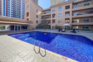 A swimming pool with a depth marker, surrounded by a building with balconies and a tiled patio area. - Scandinavian Loft Escape in JVC Dubai (Dubai)