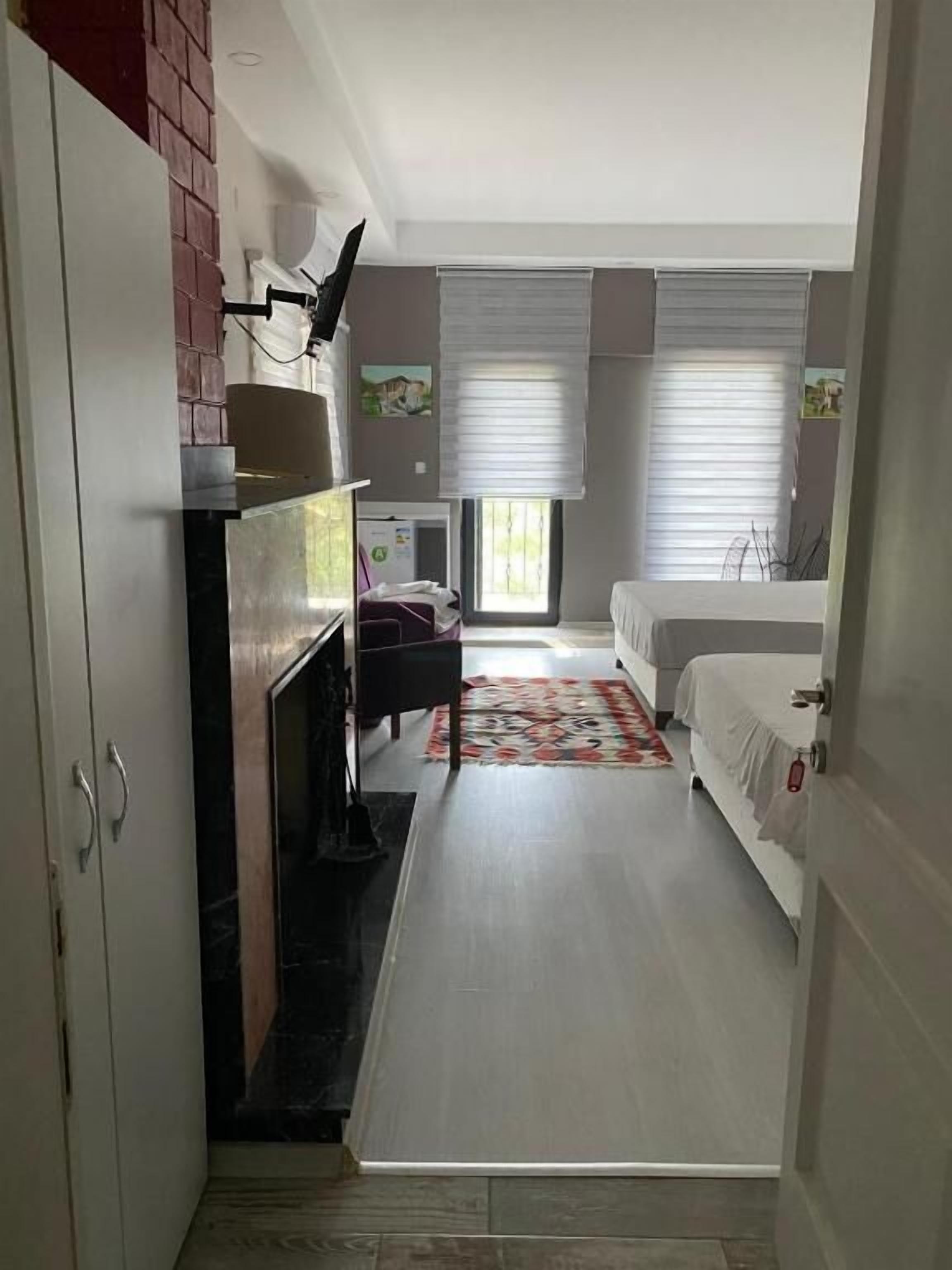 Un pasillo estrecho que conduce a un dormitorio con cama, una mesita y una silla. A la izquierda hay una chimenea y una ventana con persianas.