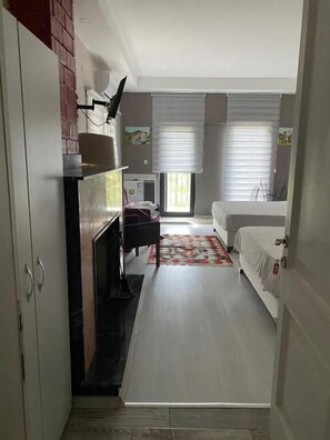 Un pasillo estrecho que conduce a un dormitorio con cama, una mesita y una silla. A la izquierda hay una chimenea y una ventana con persianas.
