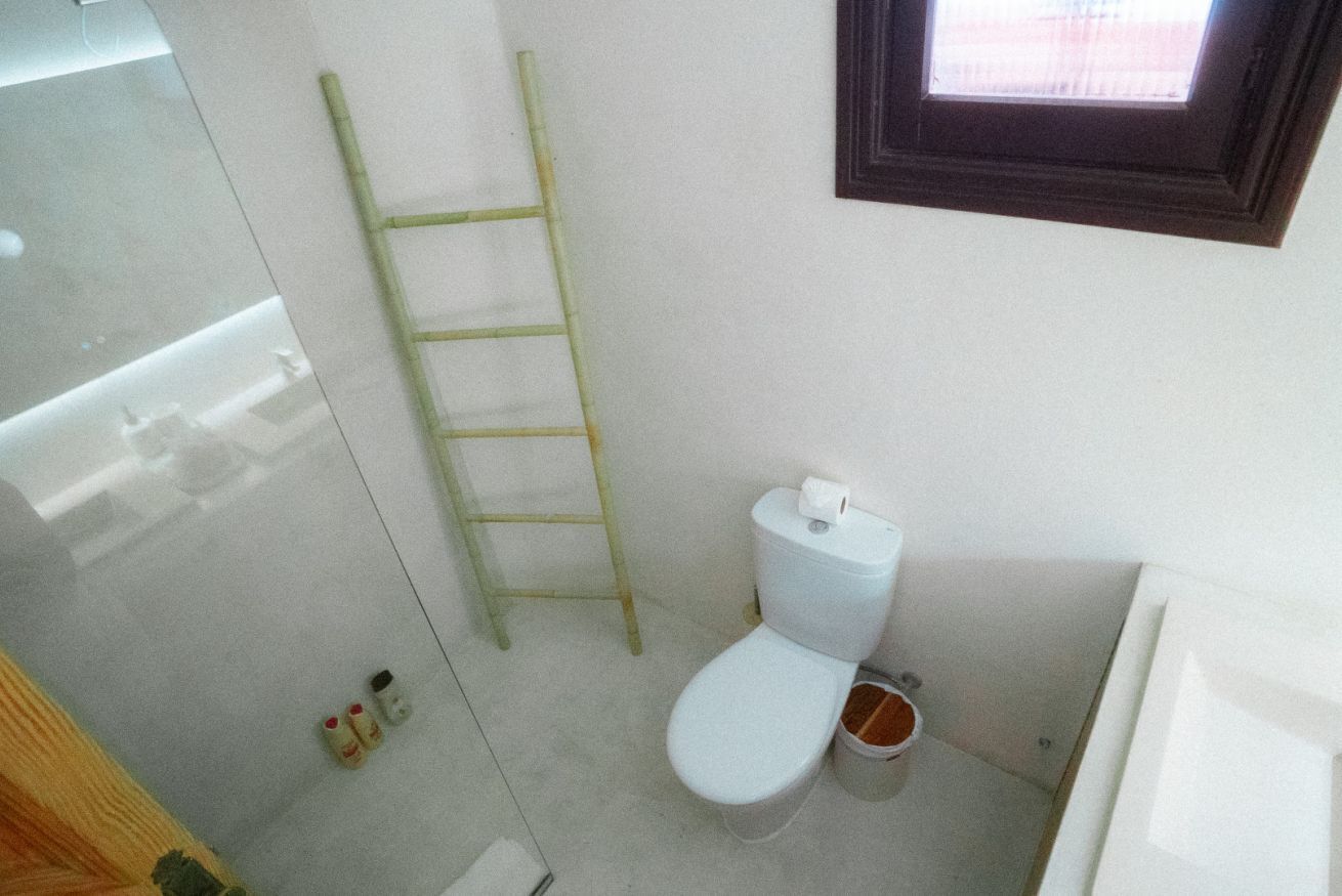A bathroom with a toilet, a small trash bin, a ladder, and a window.