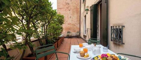A table set for breakfast with a plate of food, a glass of orange juice, and a cup, surrounded by green chairs and potted plants.