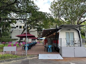 An outdoor seating area with tables and chairs, a covered seating section, and a building in the background. - Train Pod at one-north (Singapore)