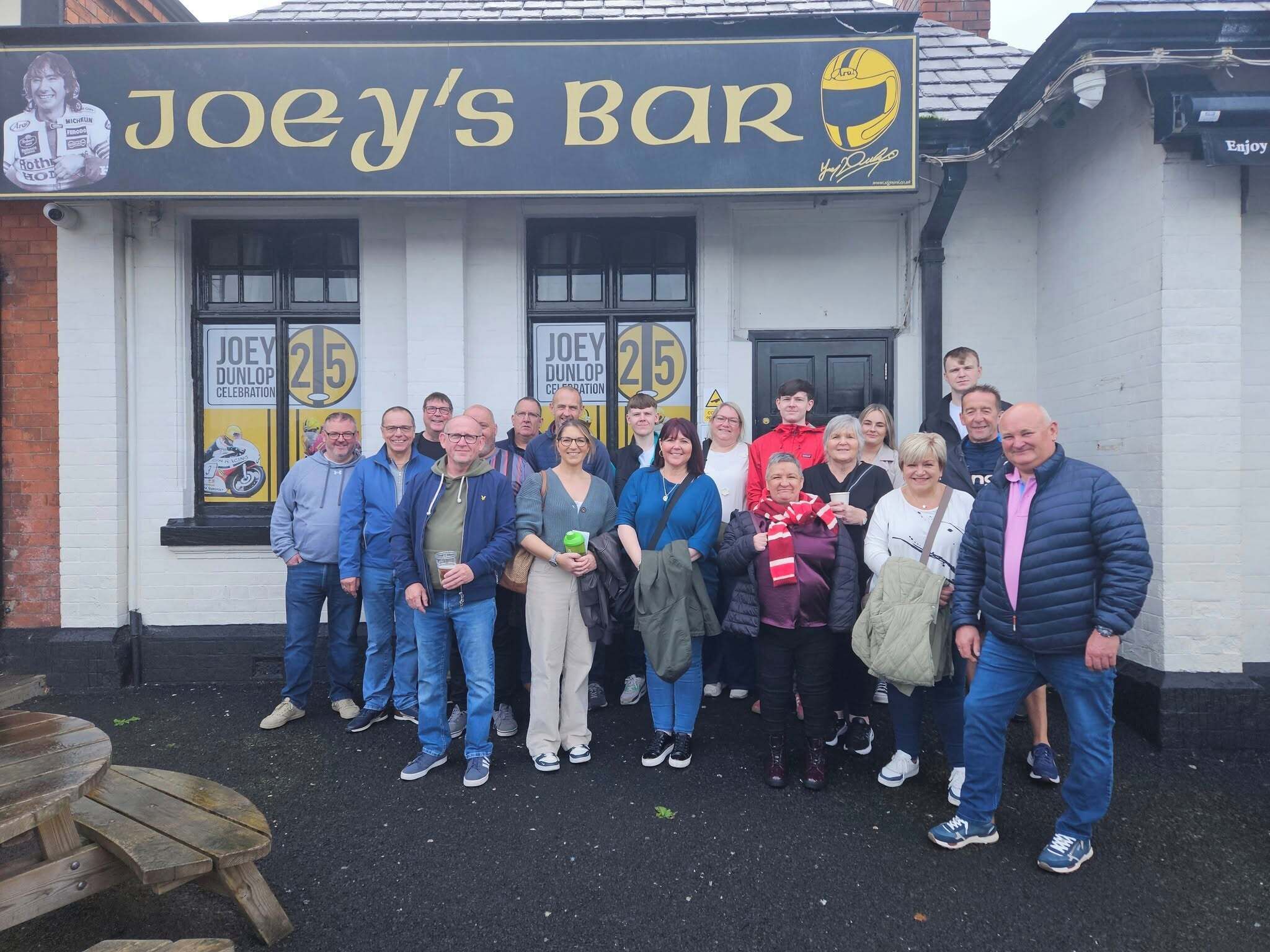 A group of people standing in front of a bar named 'Joey's Bar' with a signboard displaying 'Joey Dunlop Celebration' and the number '25'.