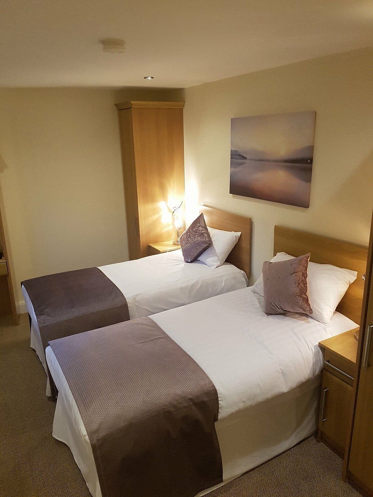 Habitación de hotel con dos camas, una mesita de noche, una lámpara, un cuadro y un armario de madera.