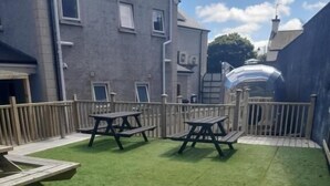 Un patio con césped artificial, mesas de picnic y un edificio de fachada gris.