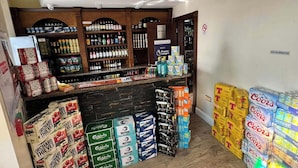 Zona de bar con estantes llenos de diferentes bebidas alcohólicas, una barra y cajas de cerveza Coors apiladas.