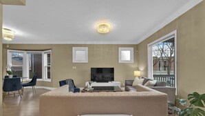 A living room with a beige sofa, a flat-screen TV, a dining area with a table and chairs, and a large window. - Olde Walkerville Wonder! (Windsor)