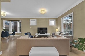 A living room with a beige sofa, a flat-screen TV, a dining area with a table and chairs, and a large window. - Olde Walkerville Wonder! (Windsor)